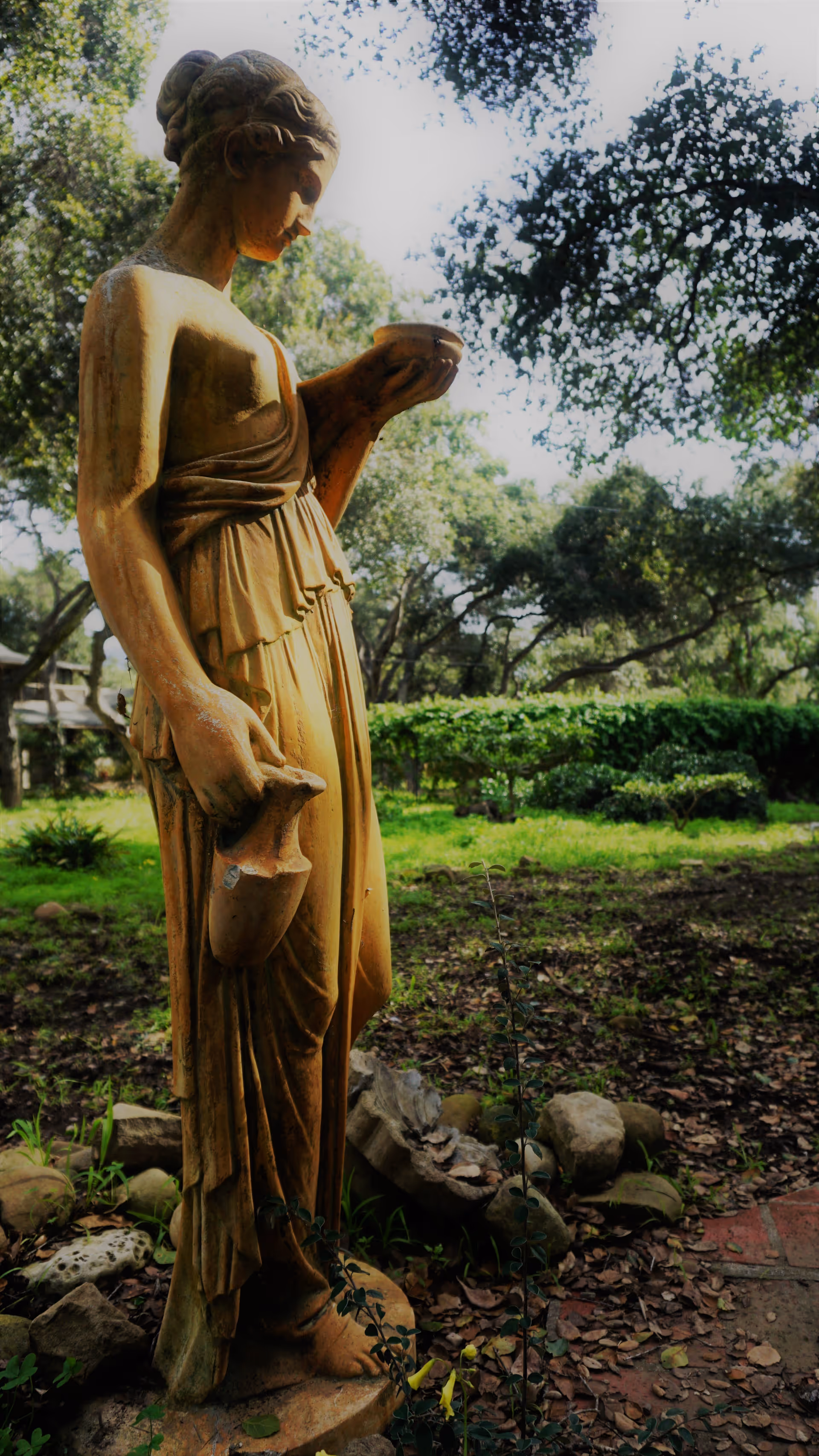Stone statue of a robed woman holding a pitcher and a bowl in a leafy garden.