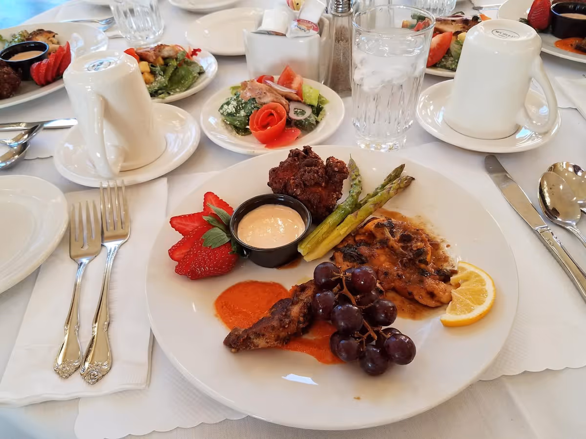 A plated meal with asparagus, grapes, strawberries, dipping sauce and a lemon wedge on a white tablecloth with utensils, coffee cups and salad plates nearby.