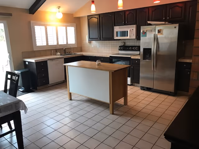 A spacious kitchen with white tiled floor and dark cabinetry. The kitchen features a central island with a wooden countertop, a stainless steel refrigerator, a microwave above a stove, a dishwasher, and a sink under a window with white shutters. Pendant lights hang from the ceiling, illuminating the area.