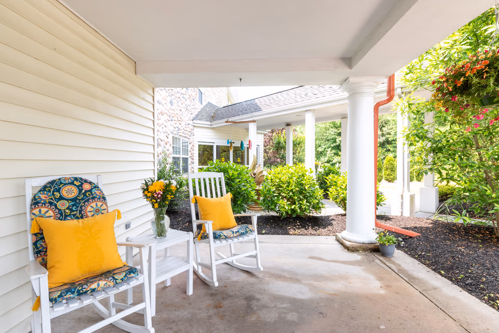Covered outdoor patio area with two white wooden rocking chairs featuring colorful cushions and yellow pillows, a small white side table with a vase of flowers, white columns supporting the roof, and green bushes and plants surrounding the space.