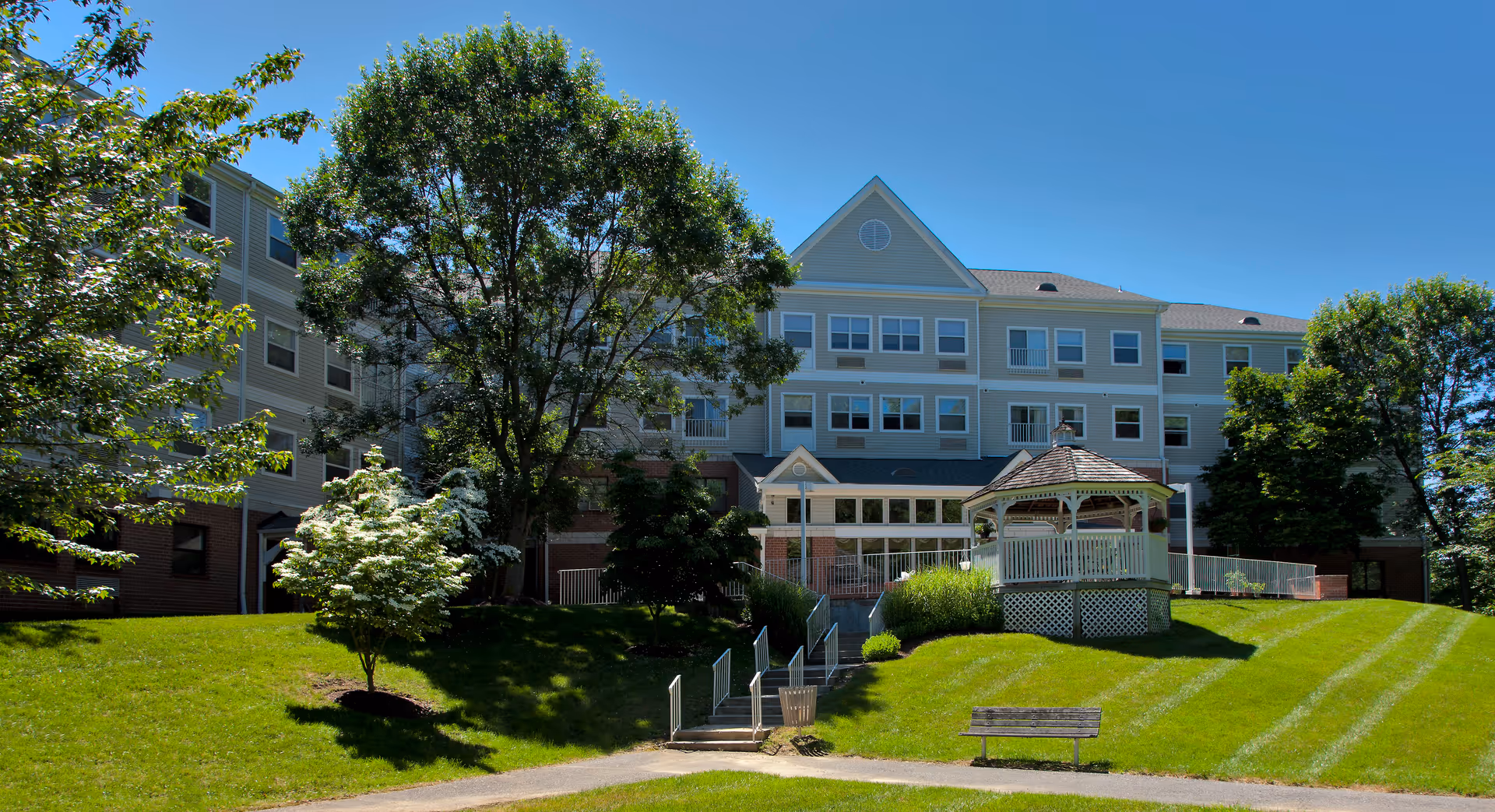 Exterior view of a senior living facility named Pin Oak Village on a sunny day. The building is three stories tall with beige siding and multiple windows. In front of the building is a well-maintained grassy area with trees, a wooden gazebo, a bench, and a paved walkway with stairs leading up to the entrance.