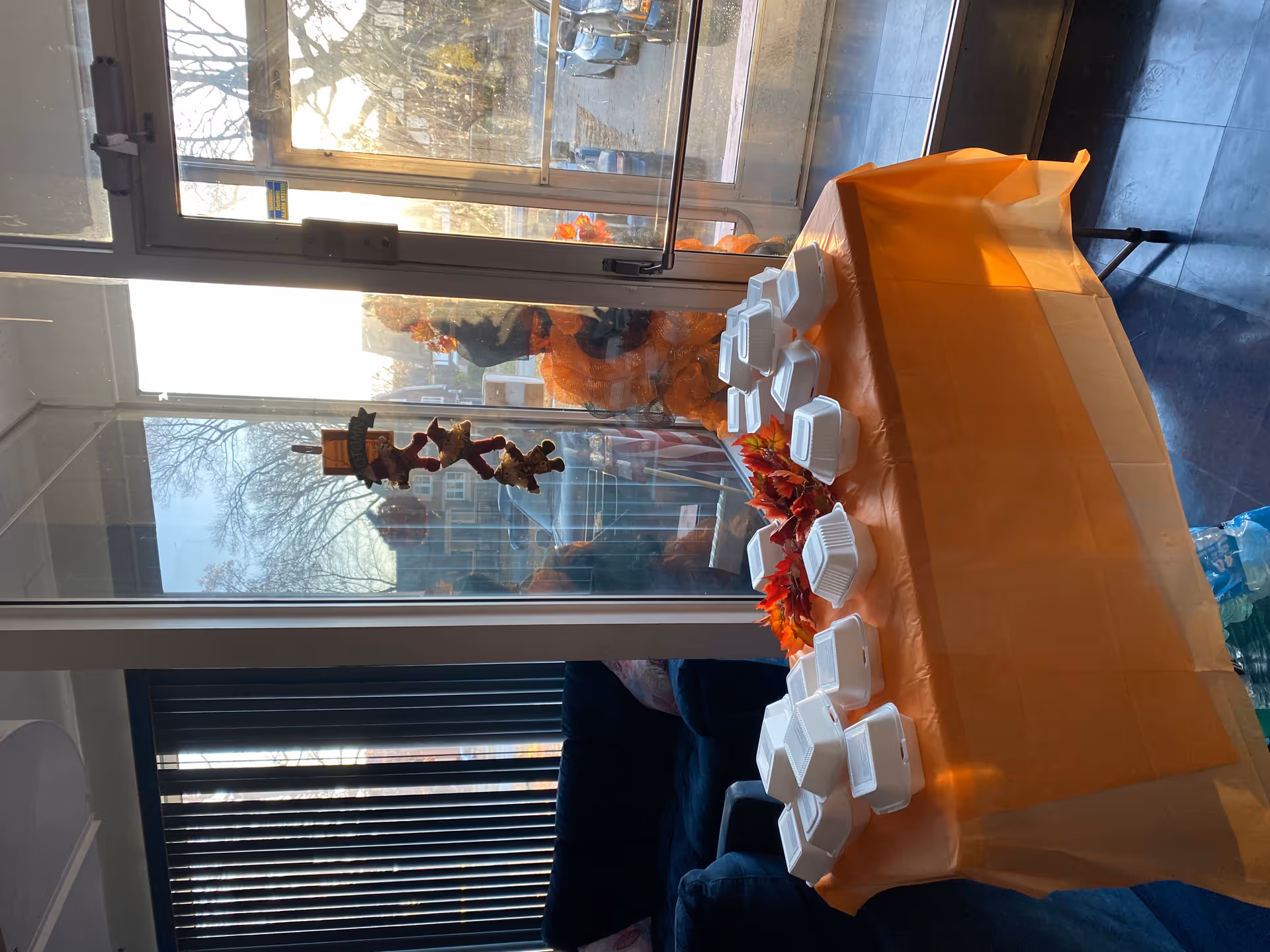 Table covered with an orange tablecloth holding multiple white takeout containers in a sunlit common room with windows and fall decorations.