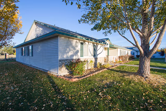 Single-story building with light-colored siding and green shutters, surrounded by grass, trees, and shrubs under a clear blue sky.