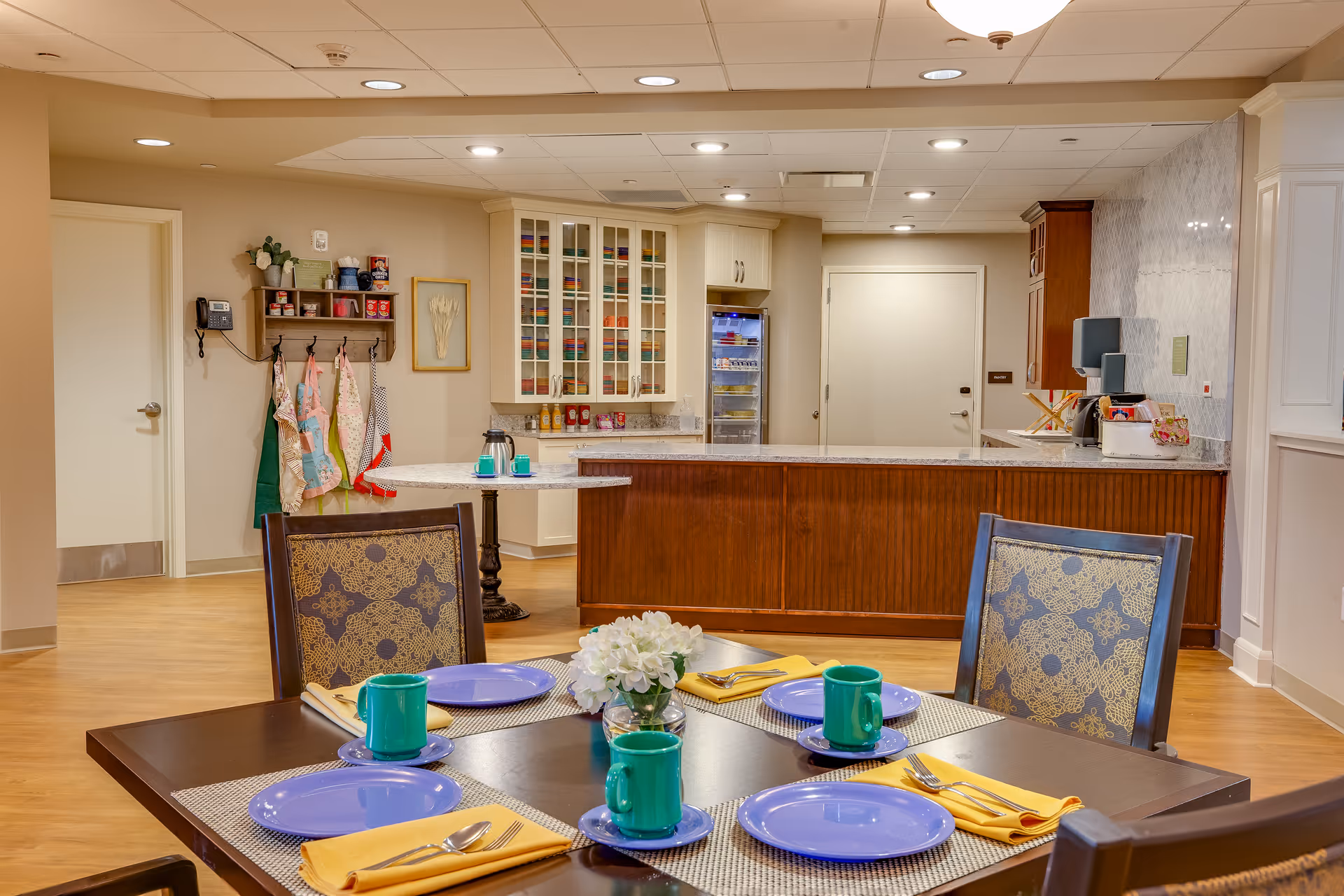 Dining area with a table set with plates, mugs, and napkins in the foreground and a serving counter and kitchenette in the background.