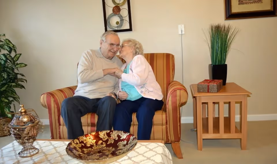 An elderly couple sitting closely on a striped armchair in a cozy living room. The woman is leaning in to kiss the man on the cheek while holding his hand. The room features a side table with a potted plant and a small gift box, a decorative bowl on a coffee table, and framed artwork on the walls.
