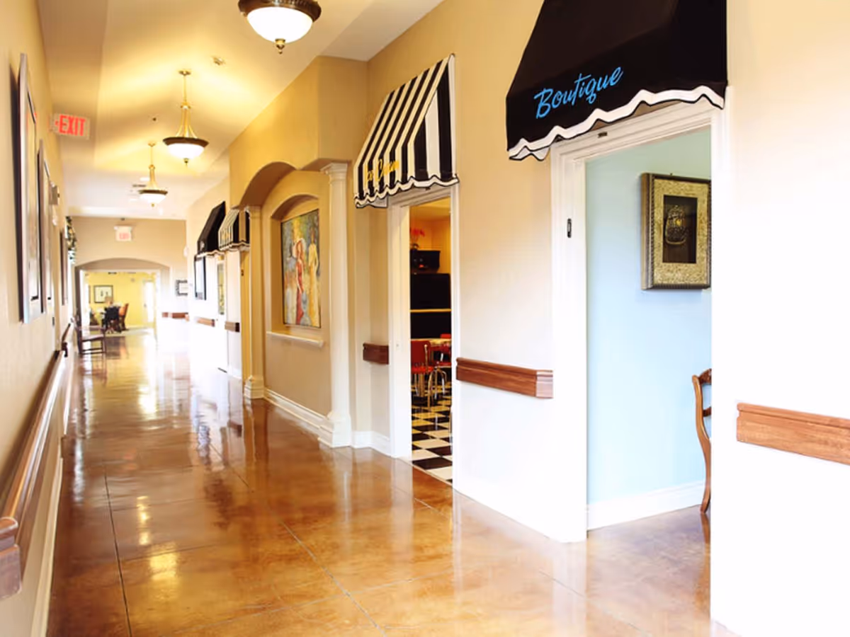 A brightly lit hallway in a senior living facility with polished brown floors, beige walls, and ceiling lights. The hallway features several doorways with black and white striped awnings labeled 'Boutique' and 'Cafe'. There are paintings on the walls and wooden handrails along the sides.