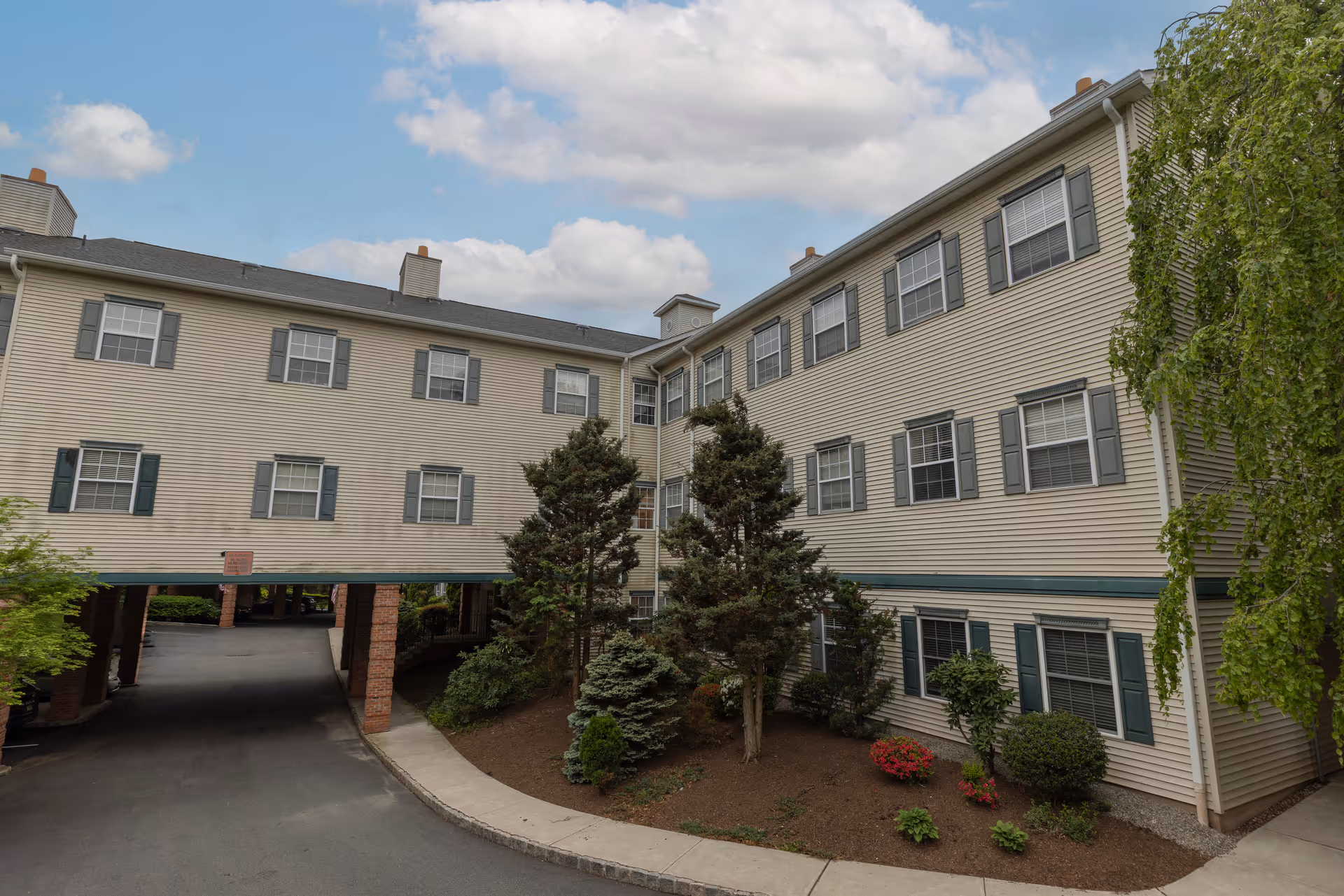 Exterior view of a multi-story residential building with beige siding and green shutters. The building has multiple windows and a covered driveway supported by brick pillars. There are trees and shrubs planted in a landscaped area near the building. The sky is partly cloudy.