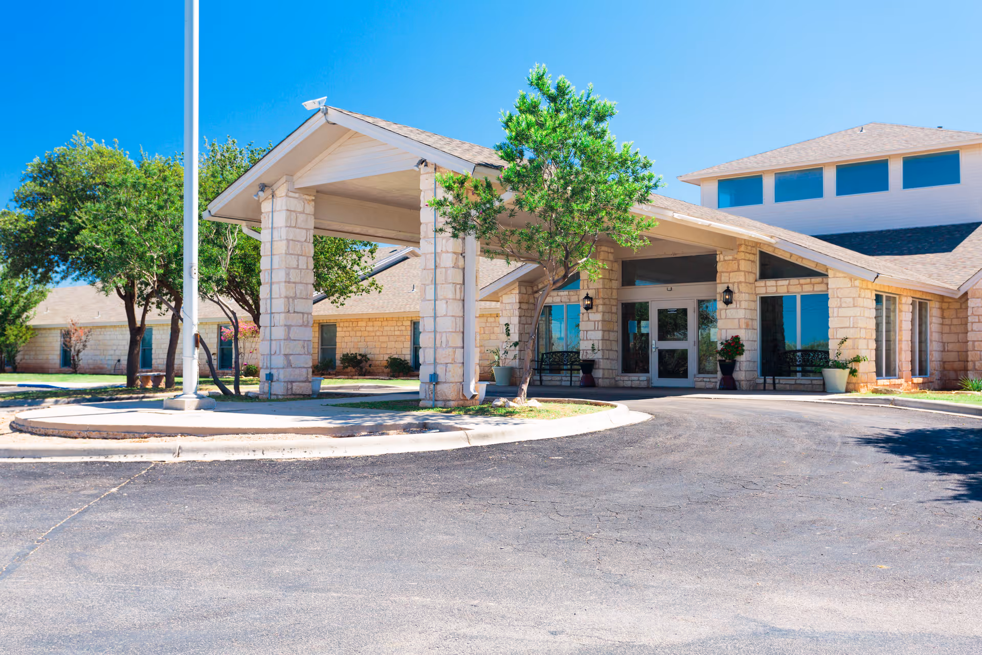 Exterior view of a senior living facility named Focused Care at Crane, showing a stone building with a covered entrance, large windows, trees, and a clear blue sky.