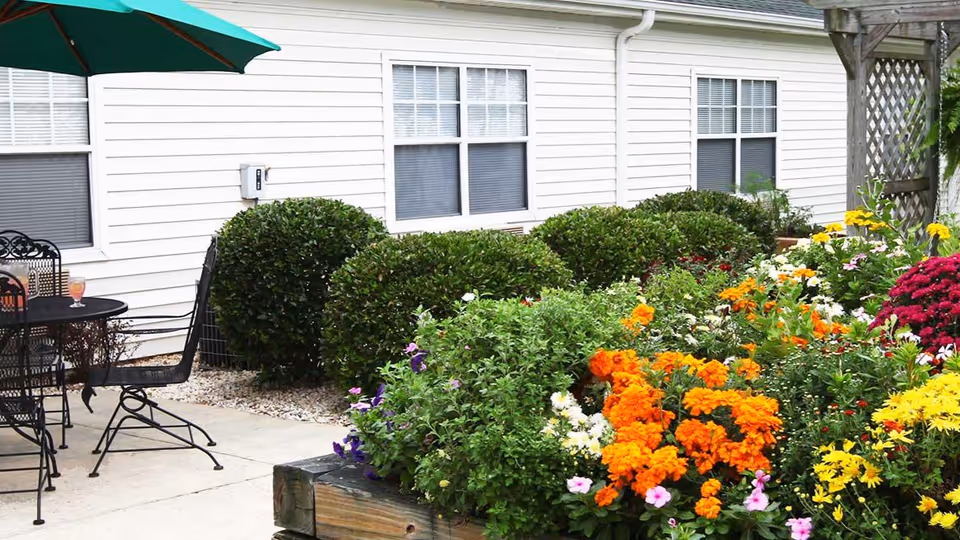Outdoor patio area with a black metal table and chairs under a green umbrella, adjacent to a garden bed filled with colorful flowers including orange, yellow, white, and purple blooms, with green bushes and a white building with windows in the background.