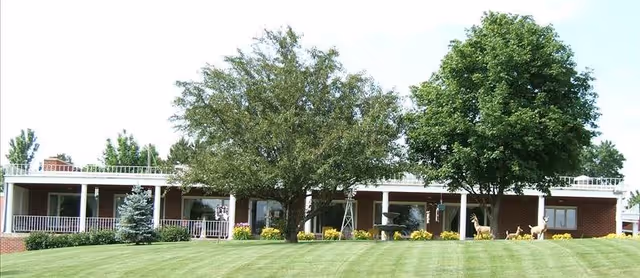 Front view of a single-story brick building with a covered porch, surrounded by green grass, trees, and decorative deer statues in the yard.