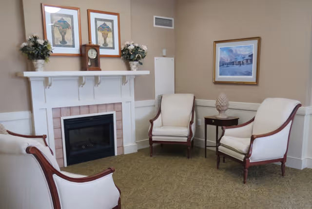 Upholstered armchairs arranged around a fireplace with framed art and floral decorations on the mantel.