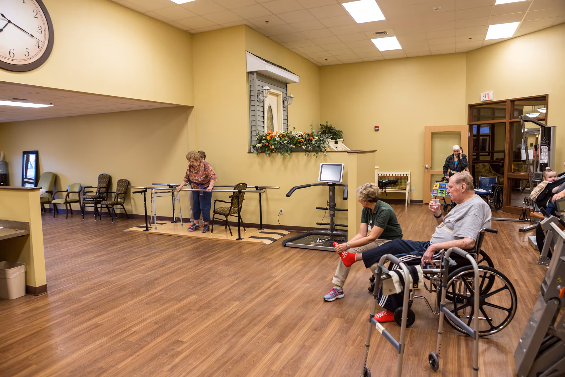 A spacious room in a senior living facility with wooden flooring and beige walls. Several elderly individuals are present, including a man in a wheelchair drinking from a cup, a woman assisting him, and another elderly woman using parallel bars for walking practice. There are chairs lined up against the walls, a large clock on the wall, and exercise equipment in the room. The setting appears to be a rehabilitation or physical therapy area.