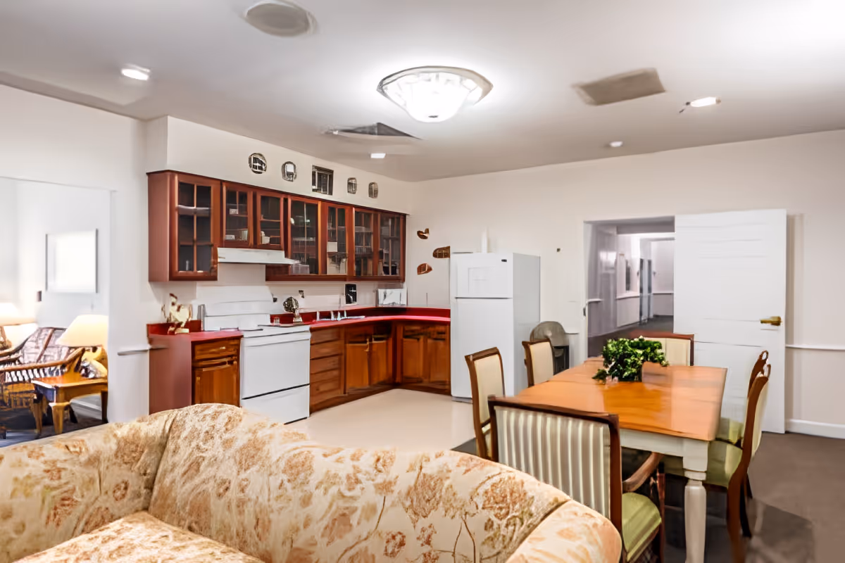 Open-plan senior living common area with a kitchen along the back wall, a wooden dining table with chairs, and a patterned sofa in the foreground.