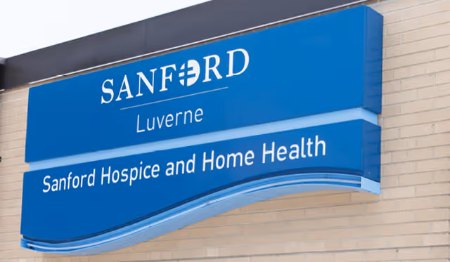 Blue sign mounted on a beige brick wall displaying the text 'SANFORD Luverne' and 'Sanford Hospice and Home Health'.