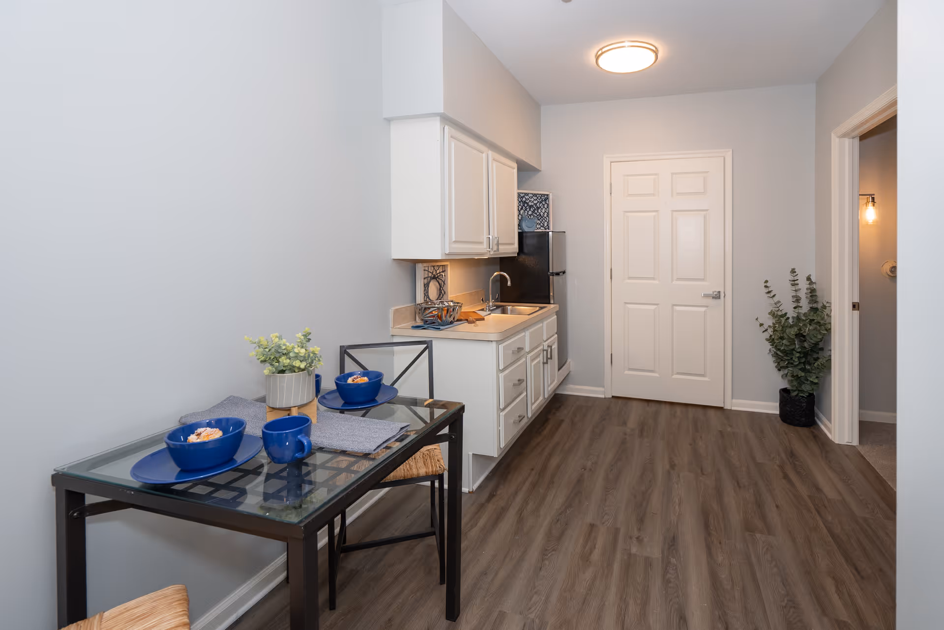 A small kitchen area with white cabinets, a sink, and a refrigerator. To the left, there is a glass-top dining table set with two blue bowls, a blue mug, and a small potted plant. The floor is wood laminate, and there is a closed white door at the end of the room. A green plant is placed in a black pot near an open doorway on the right.
