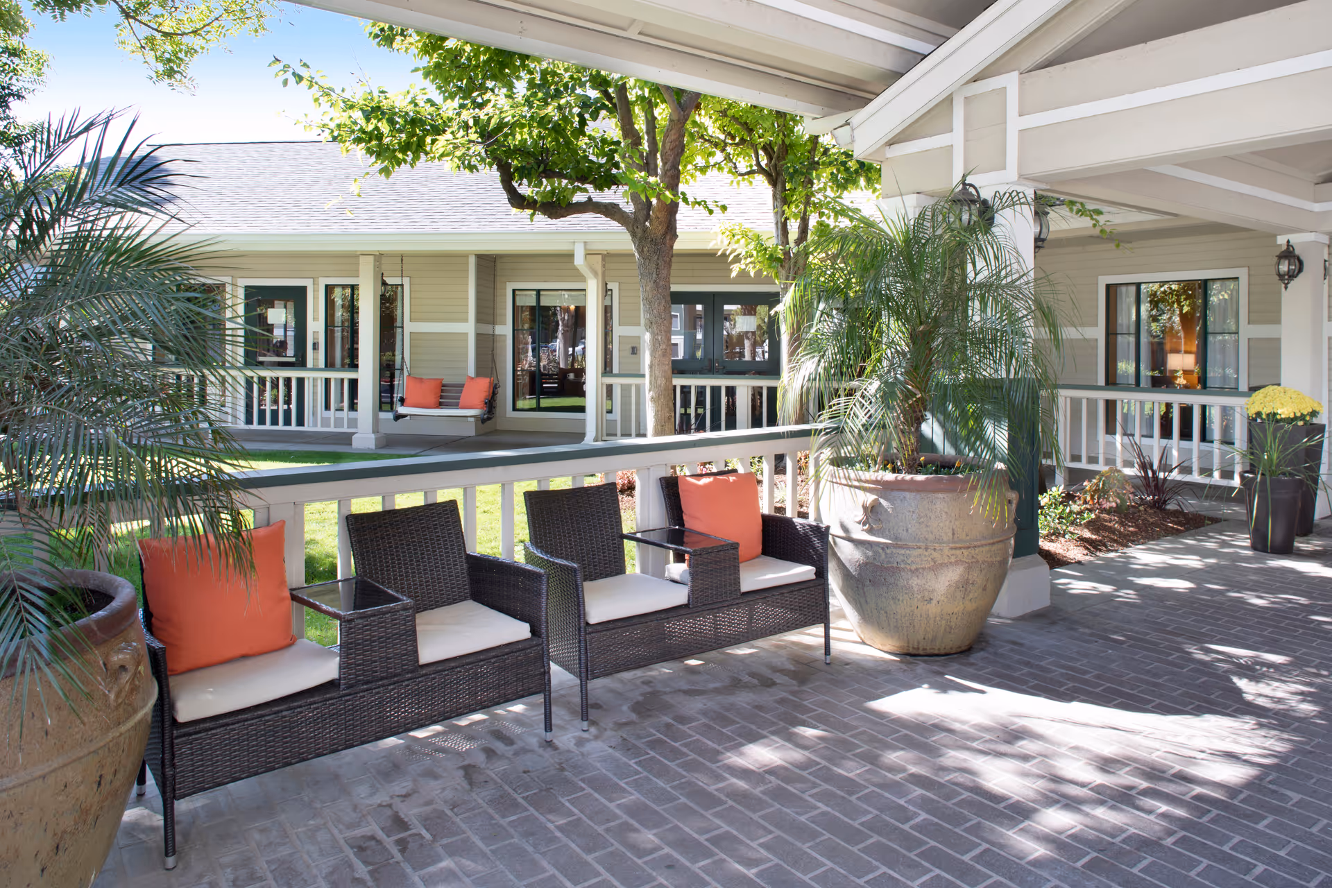 Outdoor covered patio area at Ivy Park at Salinas with wicker seating featuring white cushions and orange pillows, large potted plants, and a view of the building exterior with windows and a porch swing in the background.