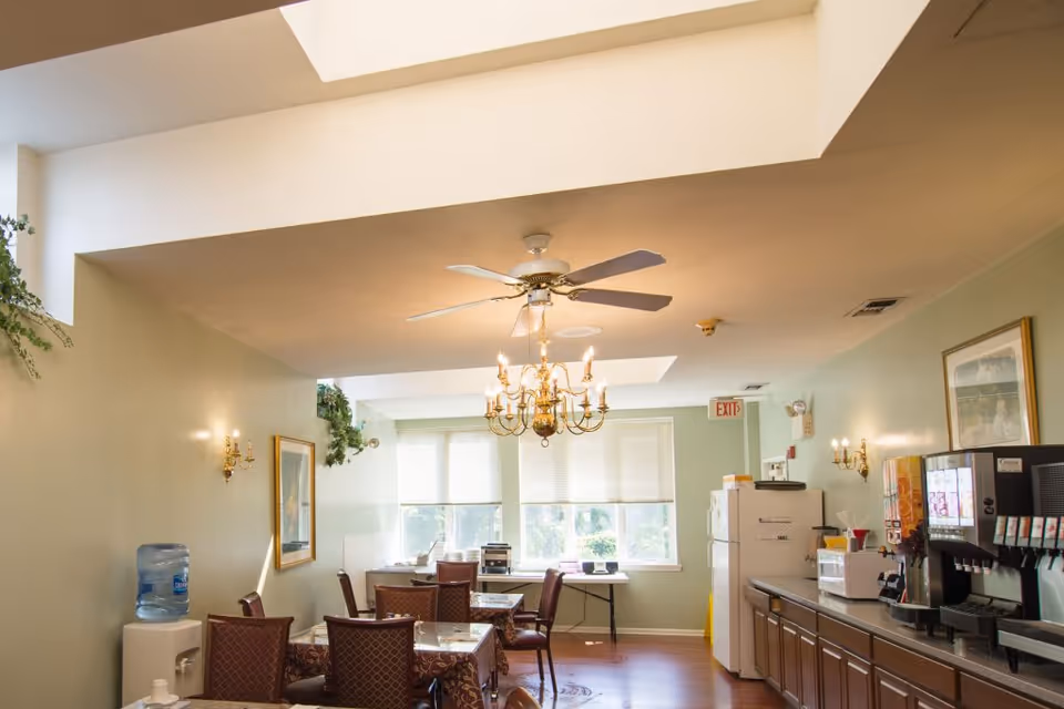 A dining area in a senior living facility with several tables and chairs, a water cooler on the left, a chandelier and ceiling fan overhead, large windows letting in natural light, and a counter on the right with a soda fountain machine, microwave, and refrigerator.