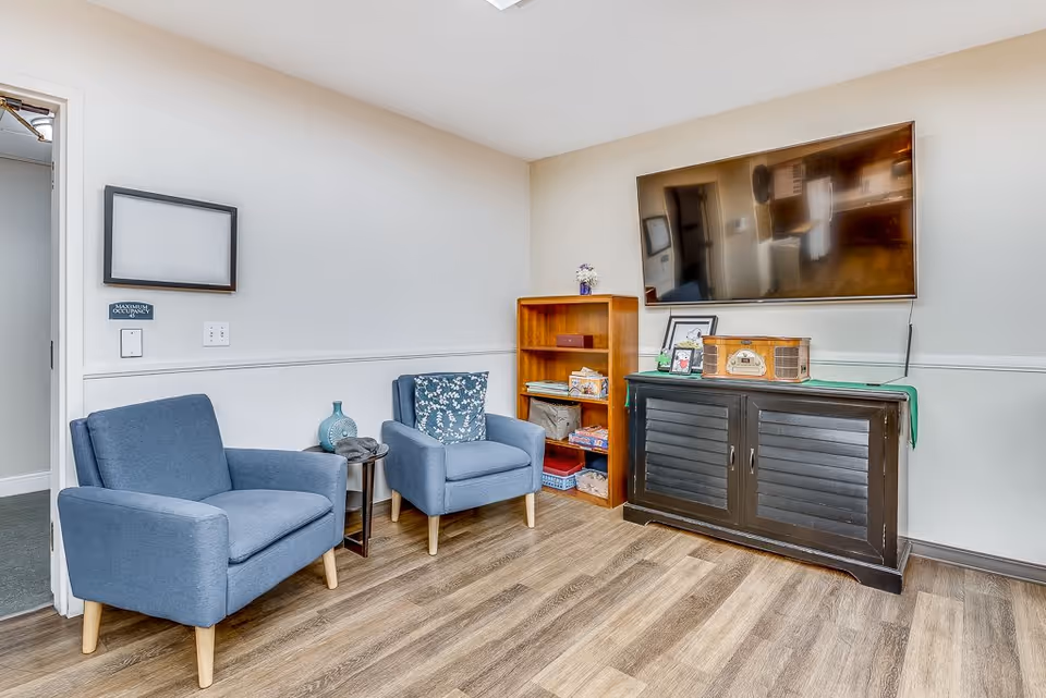 A cozy living room area with two blue armchairs, a small round side table with a decorative vase, a wooden bookshelf with various items, and a black cabinet topped with a vintage radio and framed pictures. A large flat-screen TV is mounted on the wall above the cabinet. The room has light-colored walls and wood flooring.
