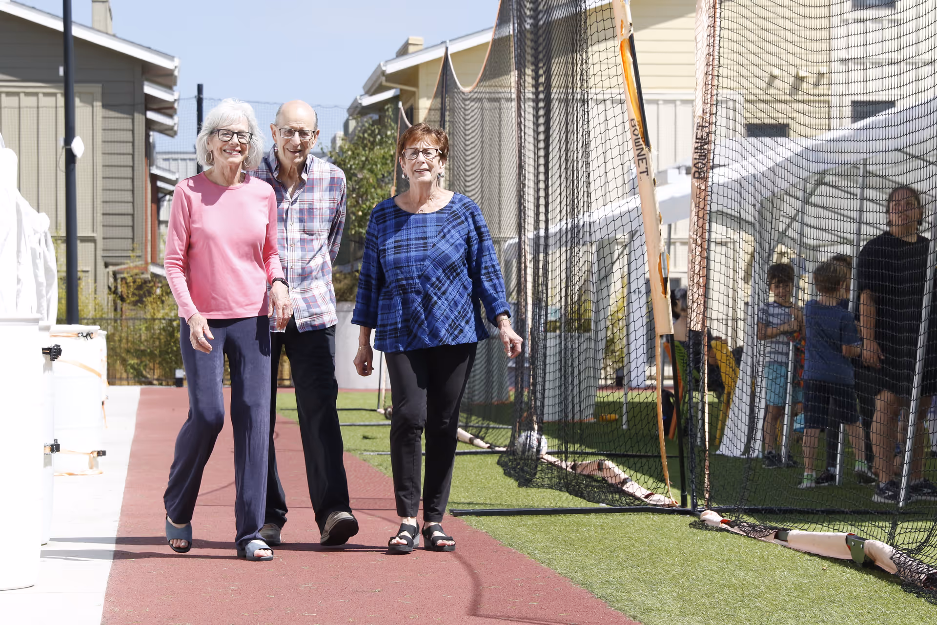 Three elderly people walking together on a red pathway outdoors near a netted area with children and an adult in the background, with residential buildings visible behind them.