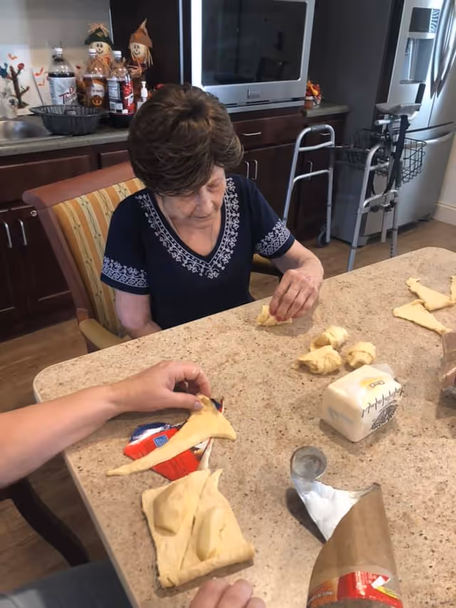 An elderly woman sitting at a kitchen table, shaping dough into crescent rolls. Another person's hands are also visible, helping with the dough. The kitchen has dark cabinets, a microwave, a refrigerator, and a walker nearby. Various kitchen items and decorations are on the counter behind them.
