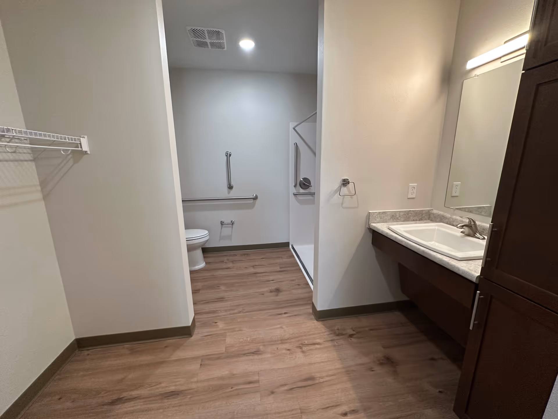 Accessible bathroom with a sink and mirror on the right, toilet and grab bars in an alcove, and wood-look flooring.