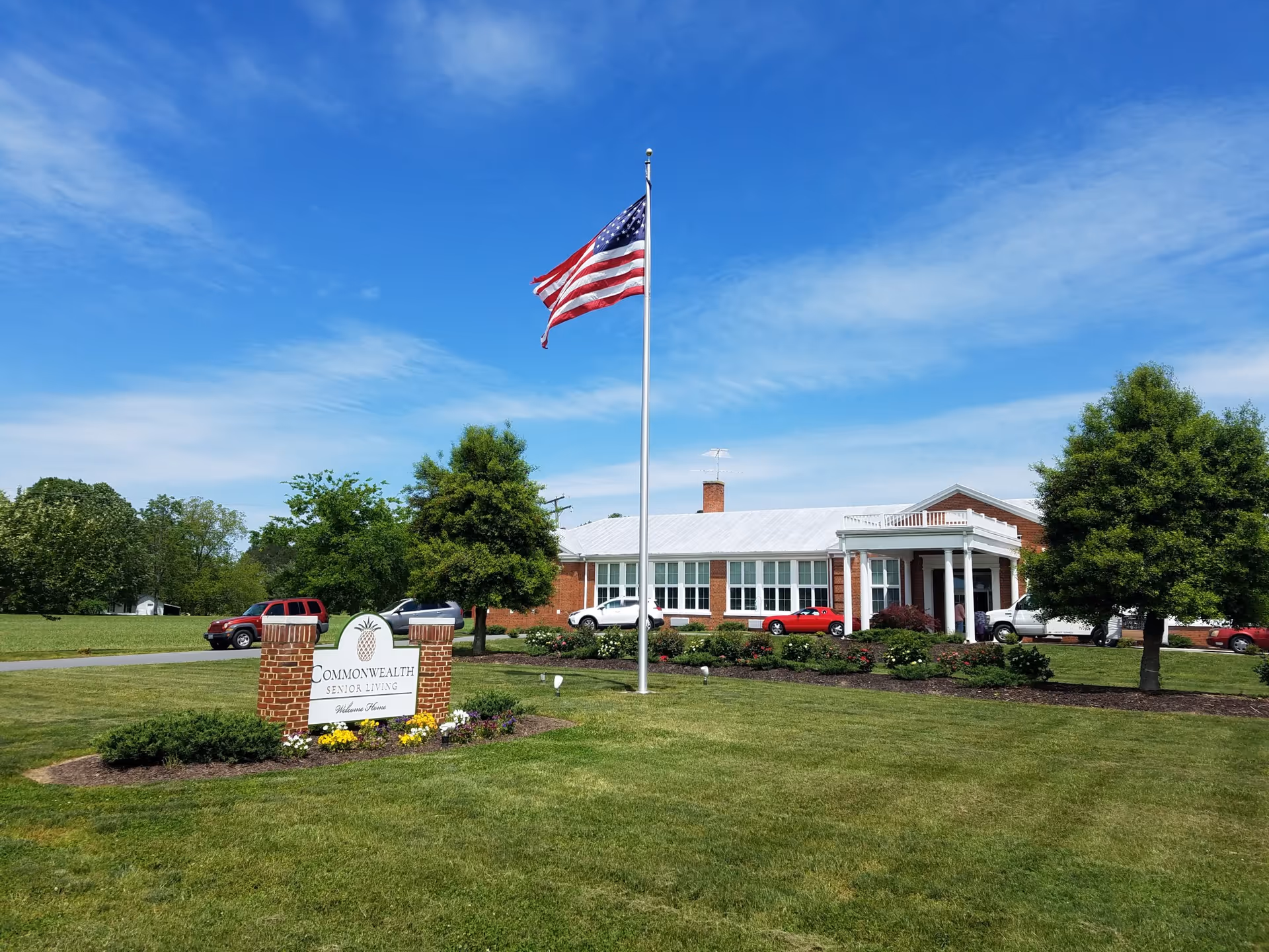 Exterior view of Commonwealth Senior Living At Farnham building with a well-maintained lawn, American flag on a flagpole, trees, and several parked cars under a blue sky with some clouds.