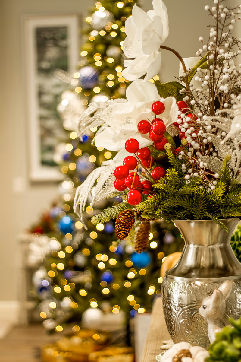Close-up of a festive holiday floral arrangement with white flowers, red berries, pine cones, and greenery in a decorative silver vase. In the background, a Christmas tree adorned with blue and silver ornaments and warm white lights is softly blurred.