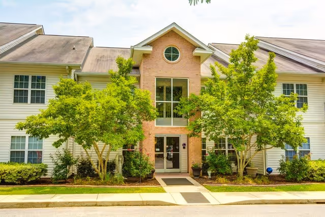 Front exterior view of a two-story senior living facility building with white siding and a central brick section featuring large windows and a glass entrance door. The building is flanked by green trees and shrubs, with a sidewalk leading to the entrance.