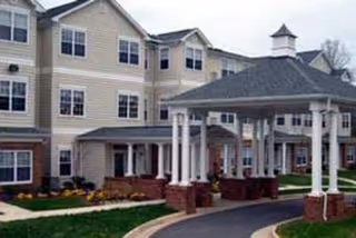 Exterior view of a multi-story senior living facility building with beige siding and multiple windows. The entrance features a covered driveway with white columns and a small cupola on top. There is a paved driveway and landscaped grass areas around the building.