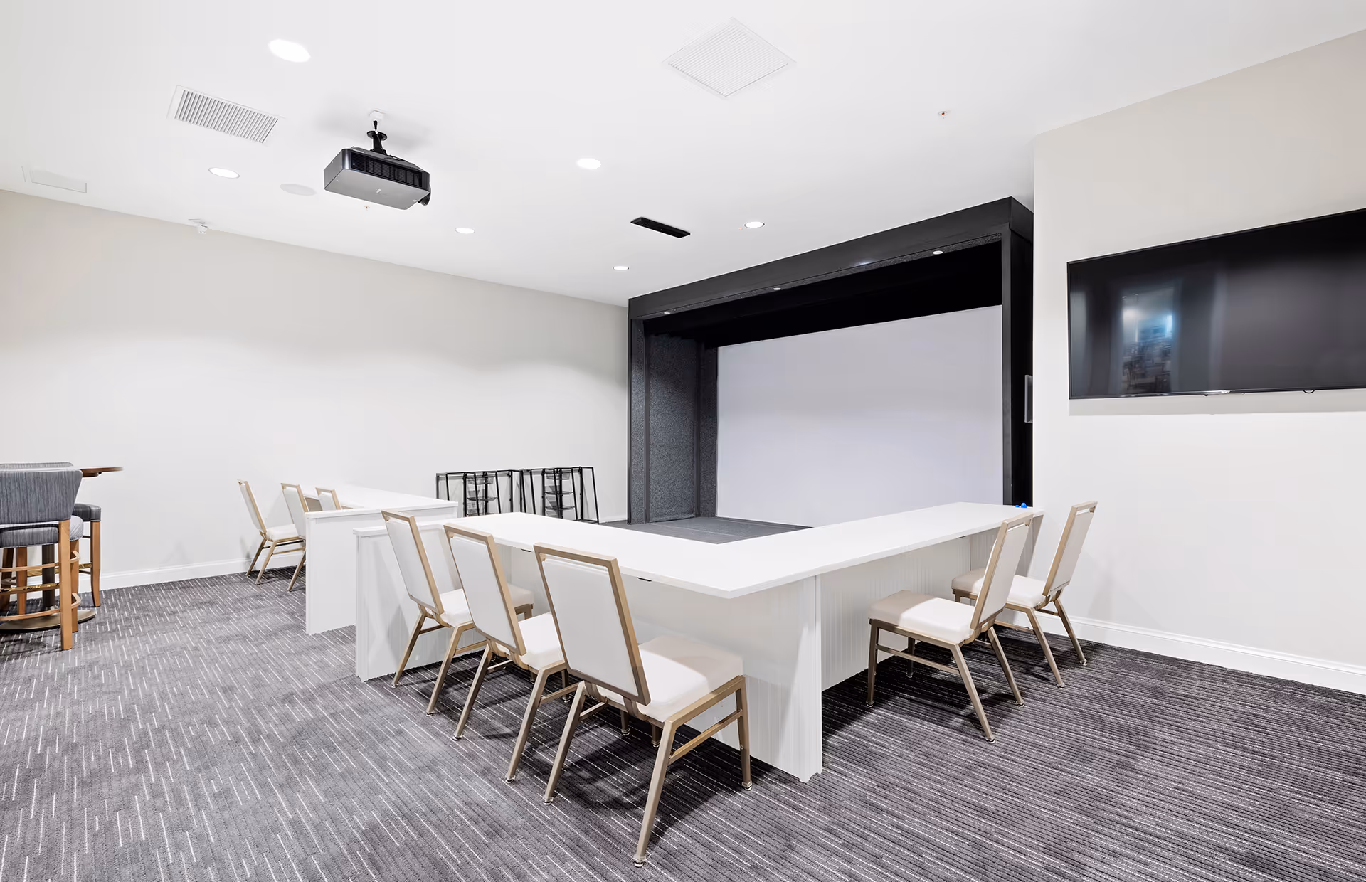 A modern conference or presentation room with a U-shaped white table surrounded by chairs. There is a large projector screen at the front, a ceiling-mounted projector, and a wall-mounted flat screen TV. The room has gray carpet and white walls with recessed lighting.