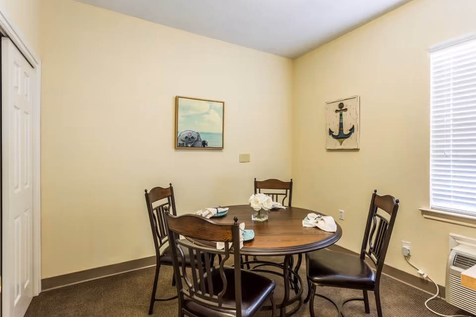A small dining area with a round wooden table and four wooden chairs with dark cushions. The table is set with folded napkins and a small vase of white flowers in the center. The room has beige walls with two framed pictures, one of an anchor and another of a seaside view. A window with white blinds is on the right side, and a white closet door is on the left.