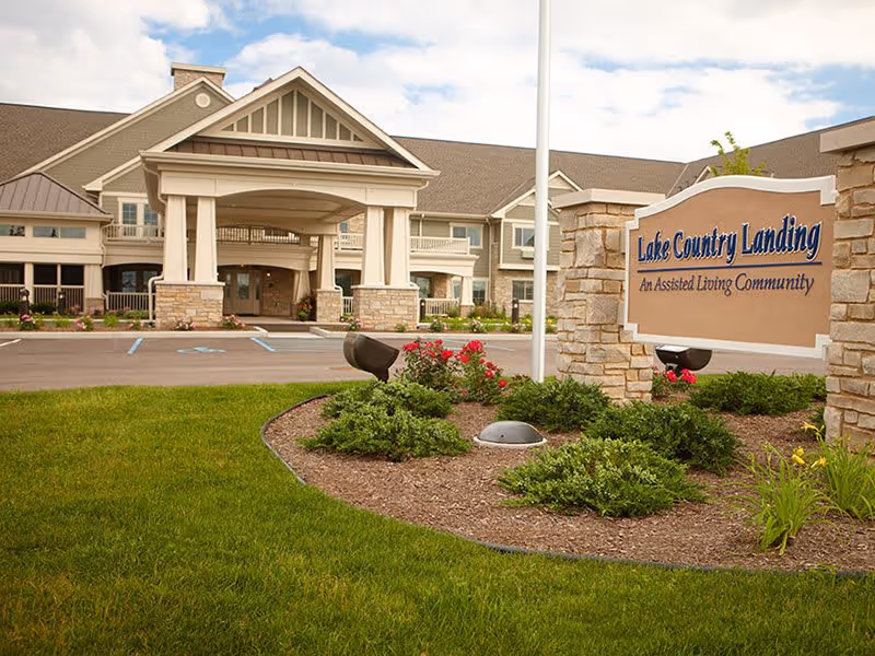 Exterior view of Lake Country Landing, an assisted living community, showing the main entrance with a covered driveway, landscaped garden with shrubs and flowers, and a sign displaying the facility's name.