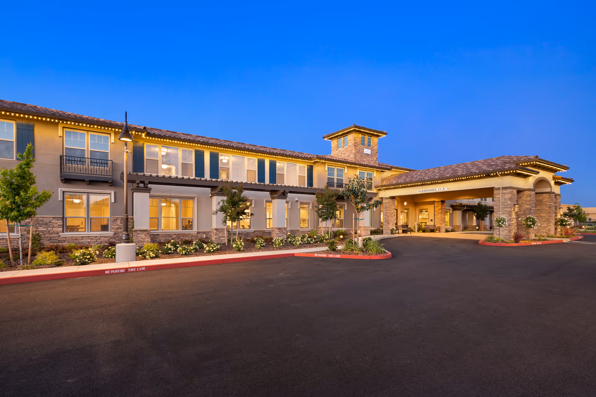 Exterior view of Oakmont of Westpark senior living facility at dusk, showing a two-story building with illuminated windows, stone and stucco facade, landscaped greenery, and a covered entrance with a clearance sign.