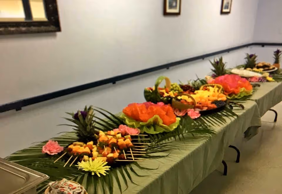 Long buffet table set up in a hallway with tropical-themed fruit platters and decorative flowers.