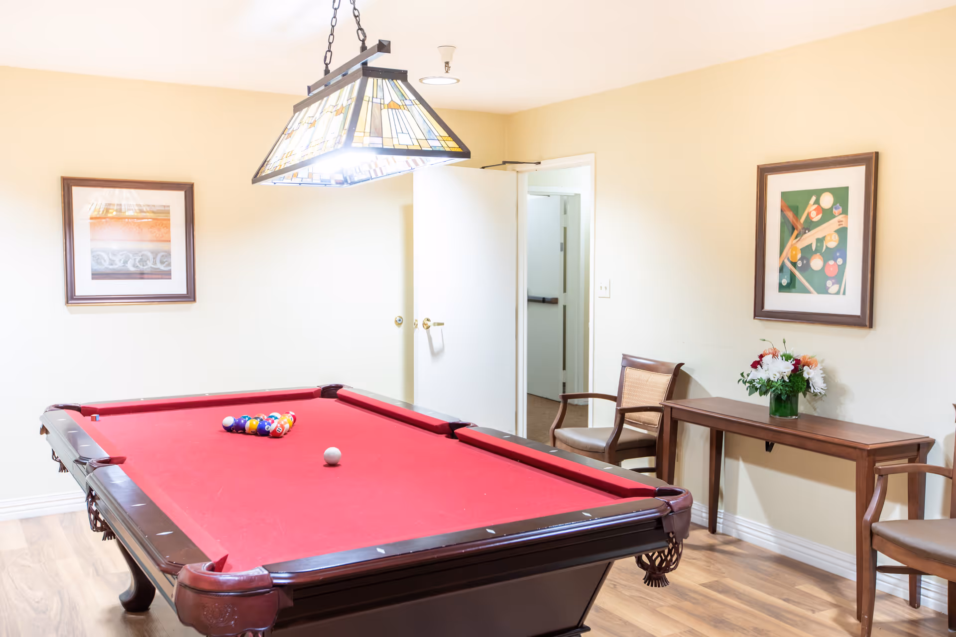 A well-lit room with a red felt pool table in the center, a hanging stained glass light fixture above it, two wooden chairs with cushions, a wooden console table with a vase of flowers, and two framed pictures on the beige walls.