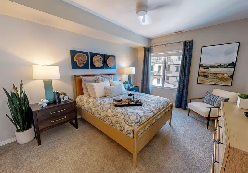 A cozy bedroom with a wooden bed featuring patterned bedding and multiple pillows. There are two bedside tables with lamps, a potted plant, and decorative items. A window with dark curtains lets in natural light, and a comfortable armchair with a striped pillow is placed near a dresser. Wall art decorates the walls, and the room has carpeted flooring.