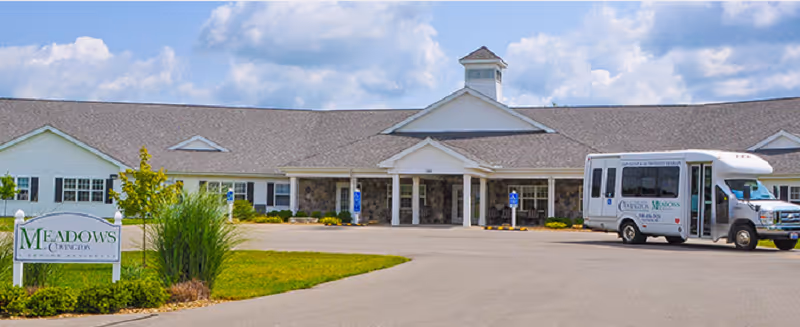 Exterior view of The Meadows at Covington senior living facility showing a single-story building with a pitched roof, a covered entrance with columns, a parking area with handicap spaces, a shuttle bus parked on the right, and a sign with the facility name on the left surrounded by landscaping.