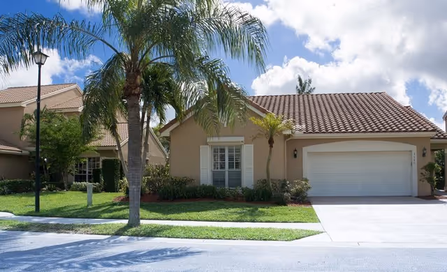 A beige suburban home with a tile roof, two-car garage, and palm trees.