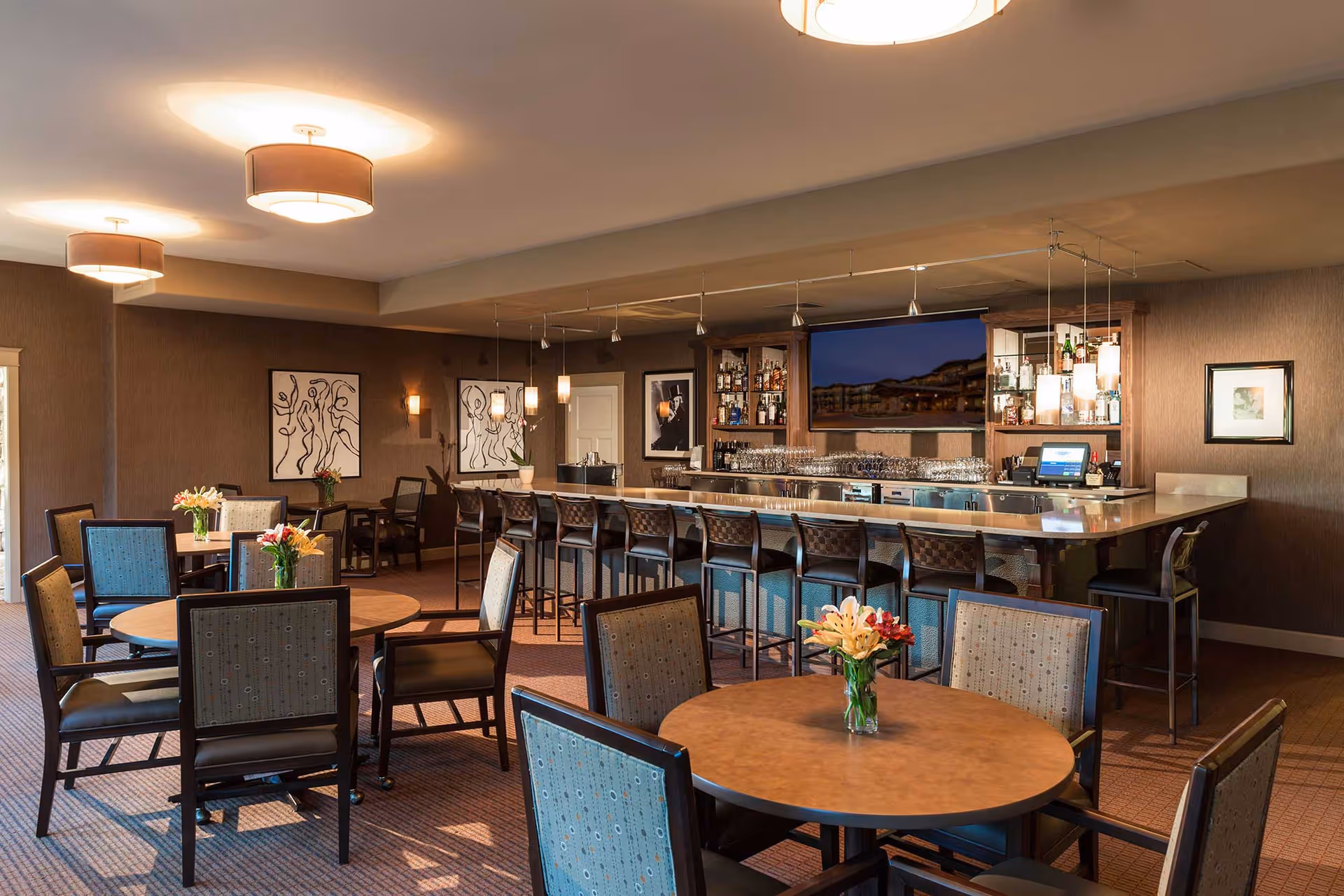 Interior view of a lounge area with round tables and chairs, each table decorated with a vase of flowers. A long bar with high stools is visible in the background, along with shelves stocked with bottles and glassware. The room has warm lighting with ceiling fixtures and pendant lights above the bar, and artwork is displayed on the walls.