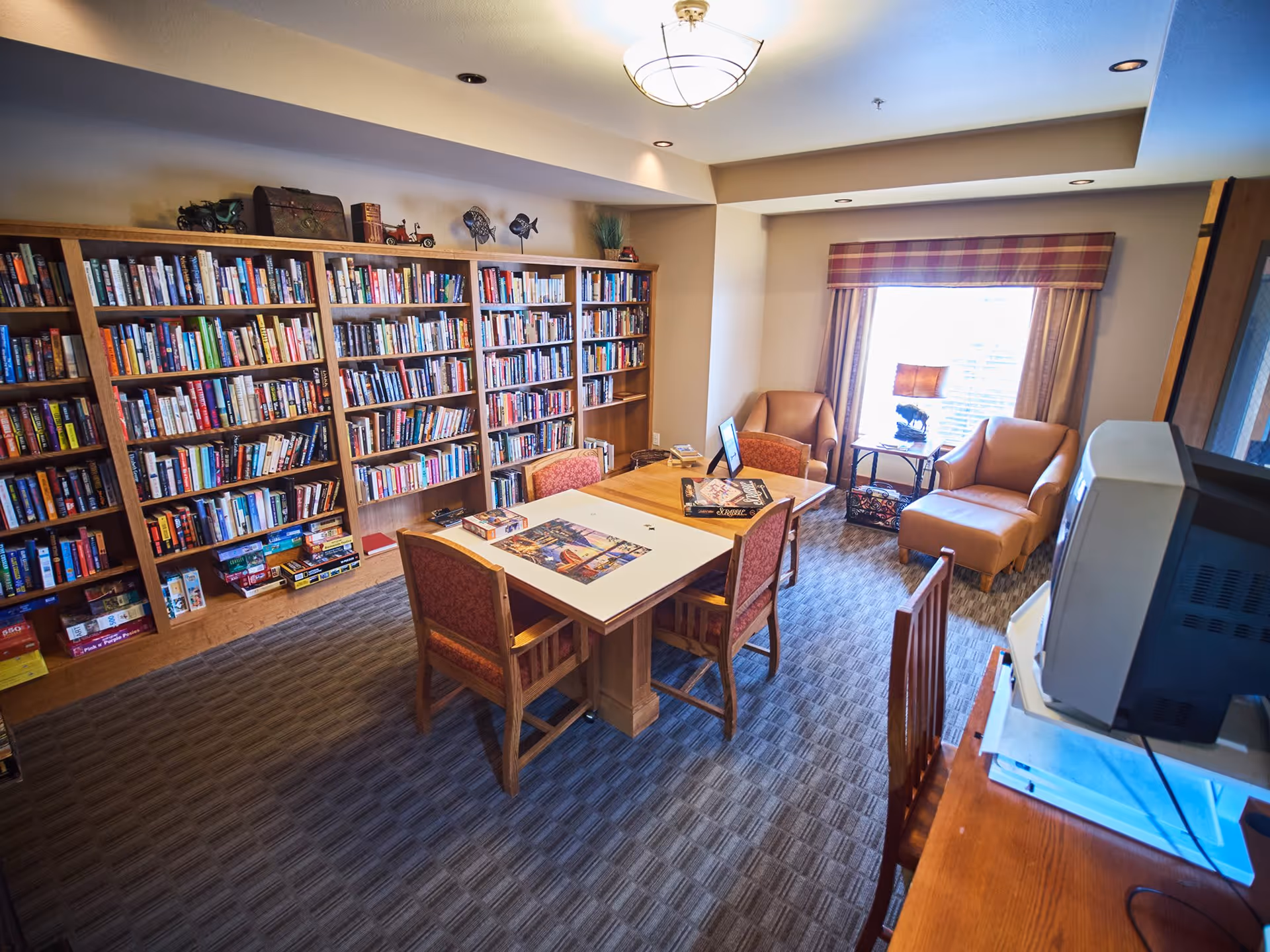 A cozy library-style common room with bookshelves lining one wall, a central table and chairs, and two armchairs by a bright window.