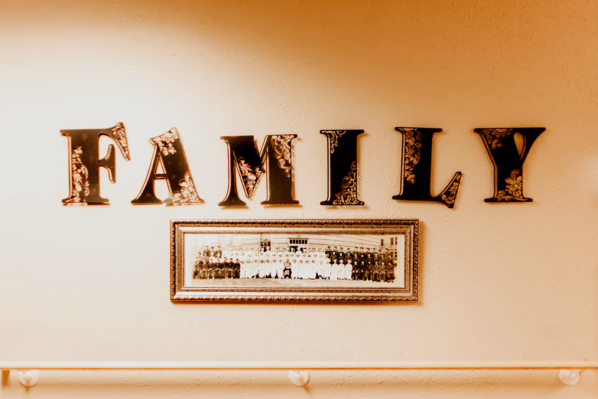 Wall decoration with large decorative letters spelling 'FAMILY' above a framed vintage black and white group photograph, mounted on a beige textured wall with a handrail below.