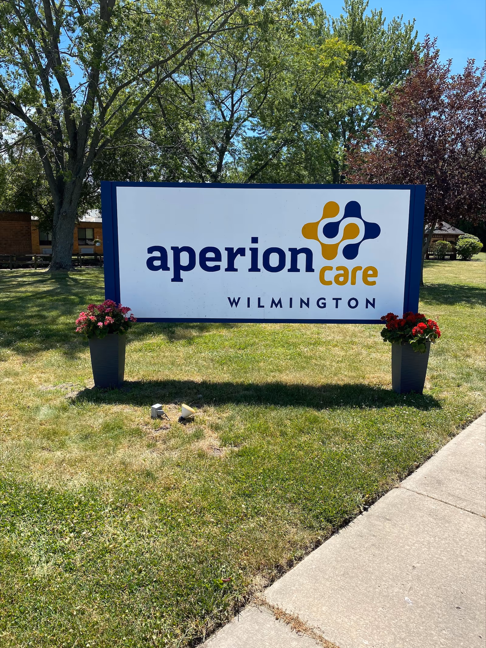 Outdoor view of a large sign for Aperion Care Wilmington placed on a grassy area with two flower pots on either side. Trees and a building are visible in the background under a clear blue sky.