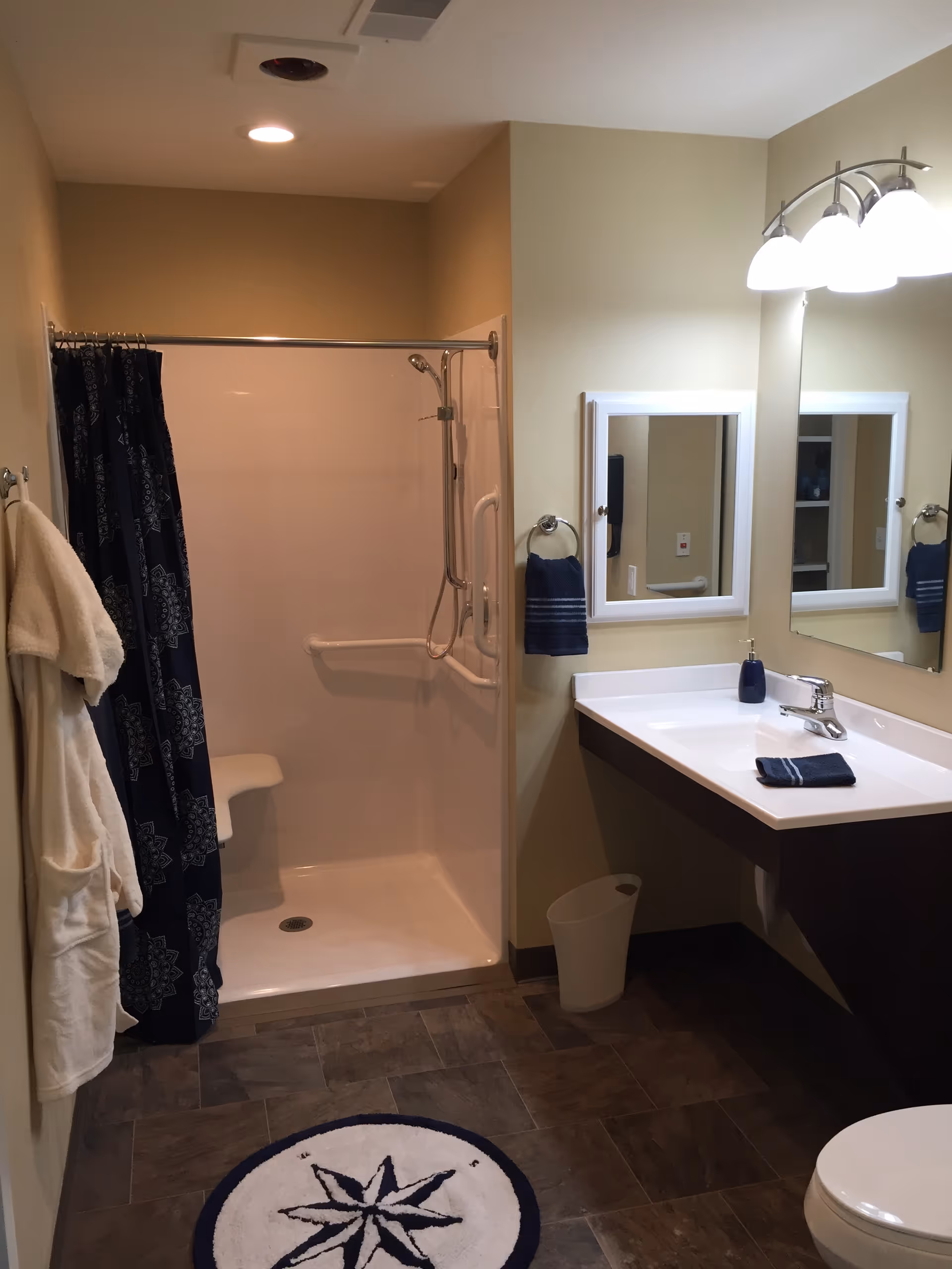 A clean bathroom featuring a walk-in shower with a built-in seat and grab bars, a dark blue patterned shower curtain, a white sink with a large mirror above it, a towel ring holding a dark blue towel, a white bathrobe hanging on the wall, a small trash bin under the sink, and a round floor mat with a compass design.