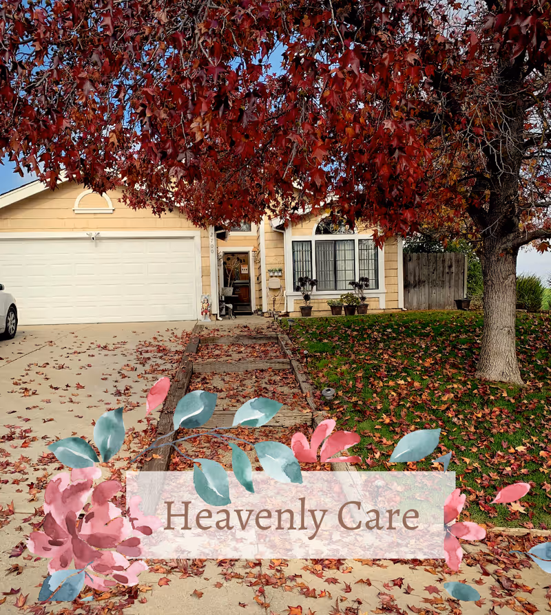 A suburban house with a beige exterior and white garage door, surrounded by autumn foliage. A large tree with red leaves stands on the right side of the driveway, which is covered with fallen leaves. There is a wooden pathway leading to the front door, and several potted plants are placed near the entrance. The sky is clear with some clouds.