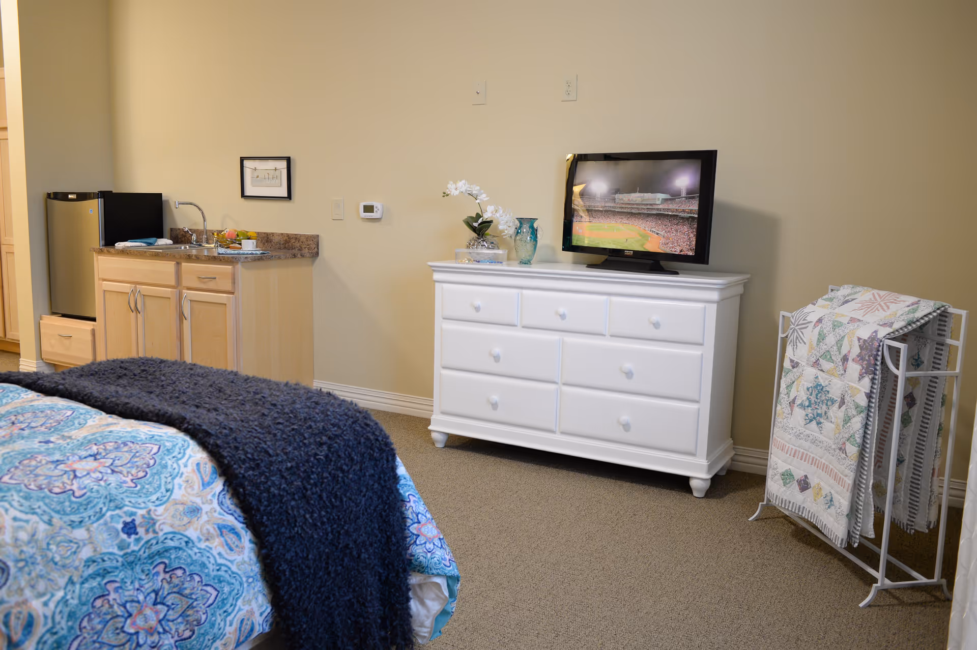 A cozy bedroom area with a bed covered in a blue patterned bedspread and a dark blue throw blanket. To the left, there is a small kitchenette with a mini fridge, sink, and countertop. On the right side, a white dresser holds a TV displaying a baseball game, along with decorative items including a vase and flowers. Next to the dresser is a white quilt rack holding colorful quilts. The walls are painted beige and the floor is carpeted.