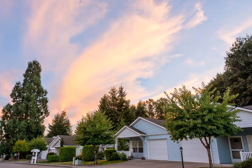 Exterior view of a senior living facility with multiple single-story houses featuring garages, surrounded by trees and greenery under a colorful sunset sky.