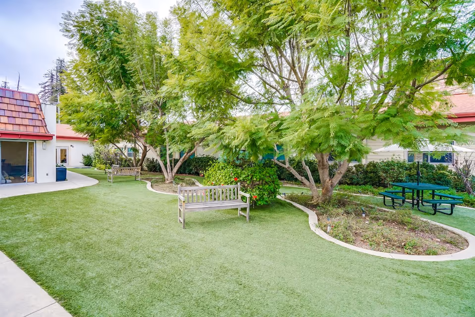Outdoor garden area at Leisure Glen Care Center with green artificial turf, several wooden benches, a green picnic table with attached benches, and trees providing shade. The surrounding buildings have white walls and red roofs.