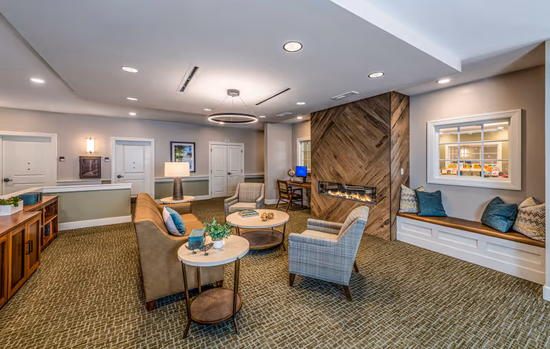 A spacious, well-lit communal lounge with sofas, armchairs, a modern fireplace, a built-in window bench, and a small desk with a computer.