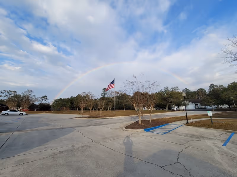 A wide concrete parking lot with a few small trees and an American flag on a flagpole in the center. There is a faint rainbow arching across a partly cloudy sky. Some buildings and trees are visible in the background.