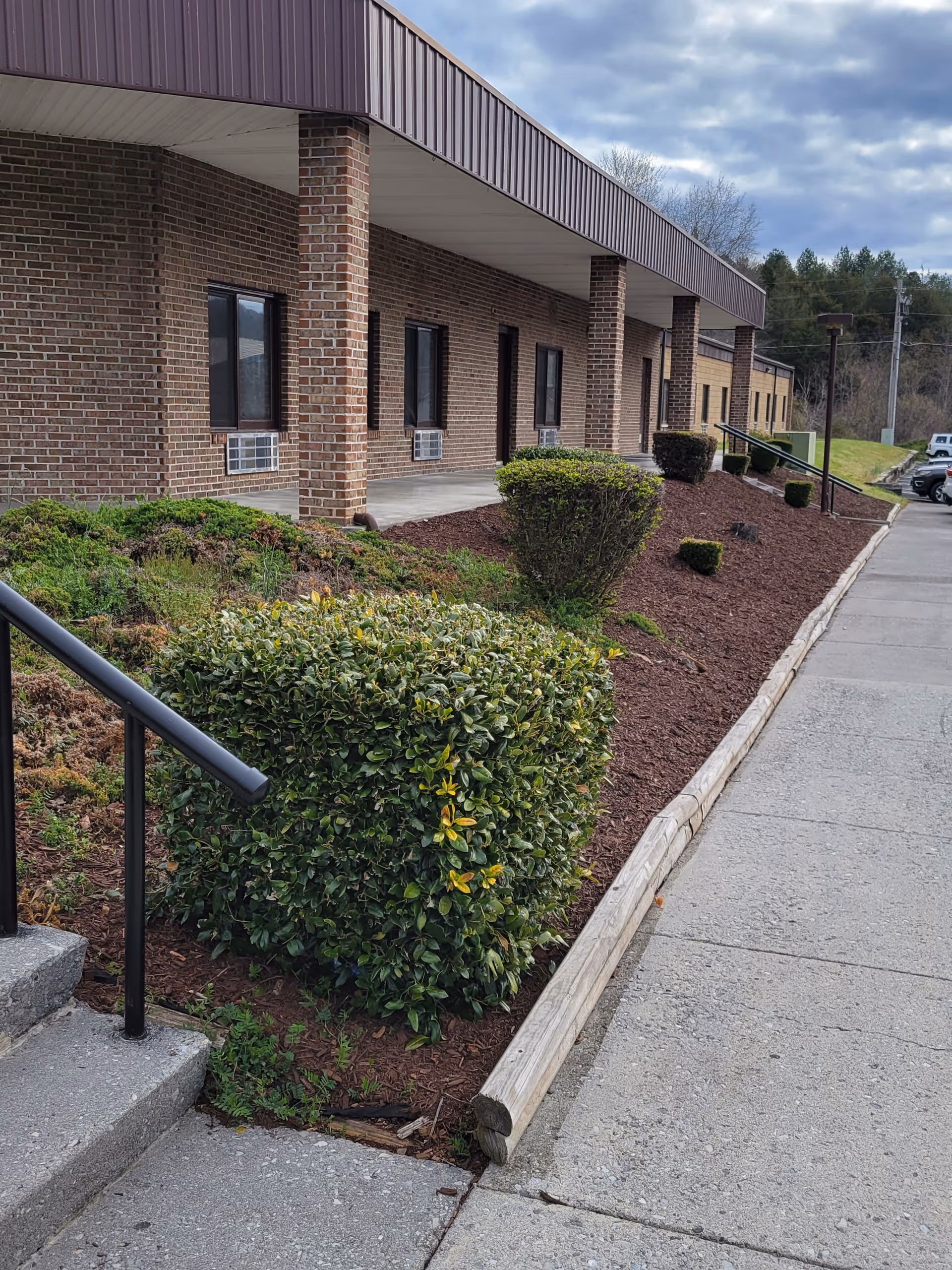 Front exterior of a brick assisted living building with a covered walkway, trimmed bushes, mulch beds, and a sidewalk.